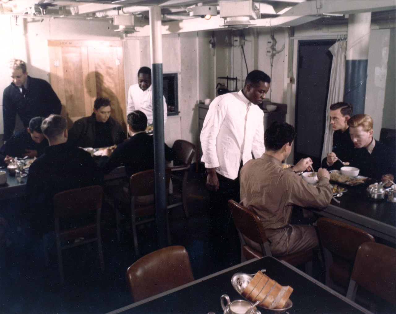 A group of men sitting at a table, enjoying a meal together, while two men stand beside the table, serving food to the group.