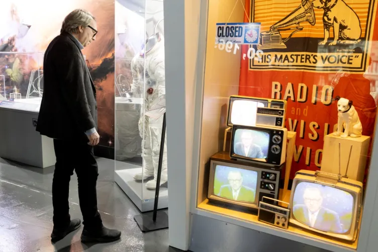 A man observing a museum display featuring a space suit and vintage televisions with a "His Master's Voice" logo and dog statue.