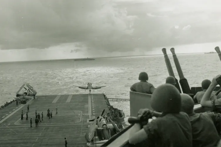 Large battleship moving through the ocean. Planes are lined up on the flight deck, with crew members bustling around.