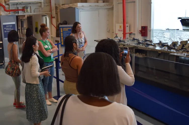 A group of women gathered around, looking closely at a detailed LEGO model of the Intrepid aircraft carrier on display at the Intrepid Museum