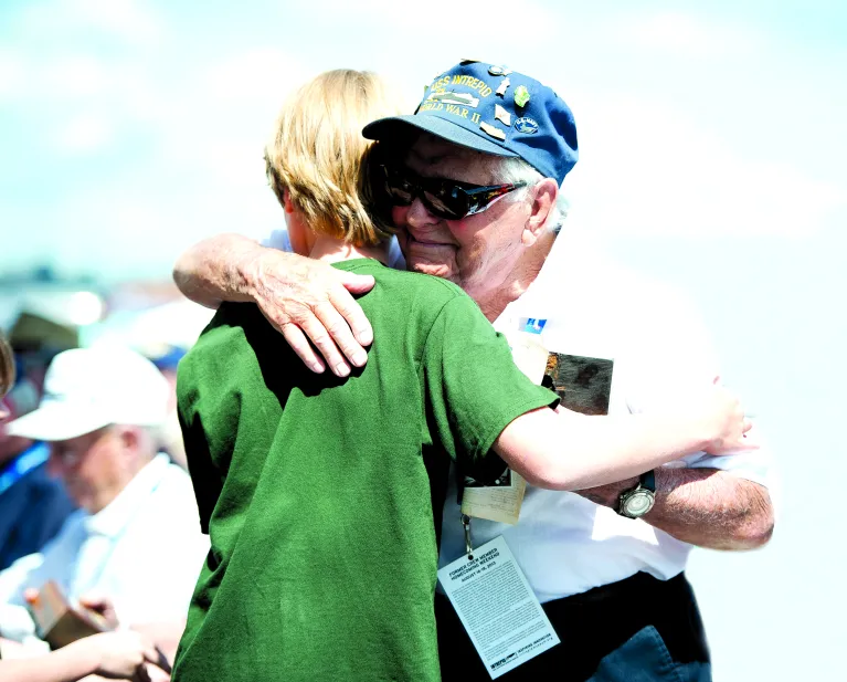 A group of veterans and their family members sit on the ship's flight deck. One veteran hugs a young child.