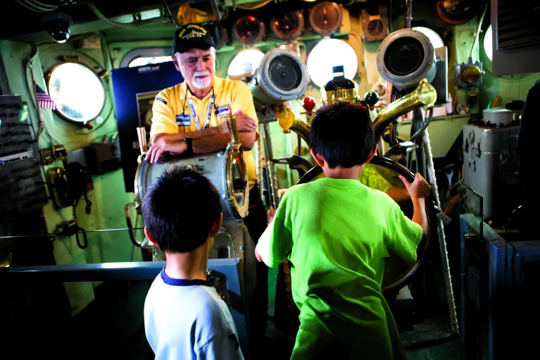 A veteran and two children are in the helm and one child is holding the steering wheel.