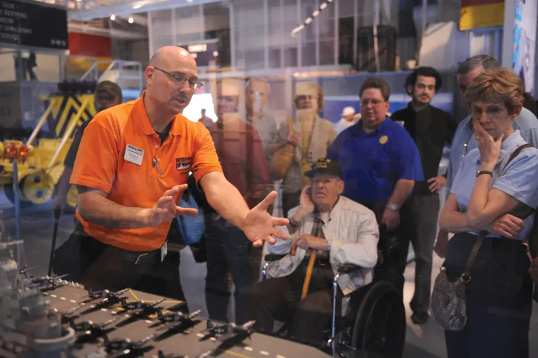 A tour guide is speaking to a group of visitors in front of a model of Intrepid.