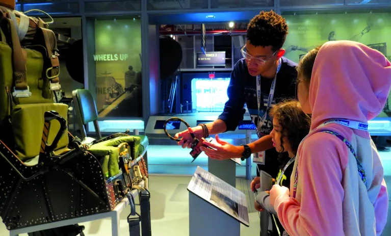 An educator is holding an artifact in front of an airplane seat and showing it to two children.