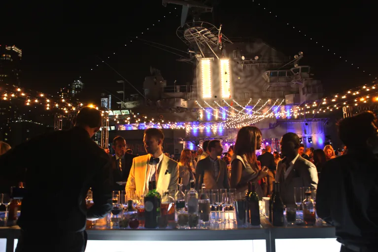 Partygoers approach a bar situated on Intrepid's flight deck at night, flanked by twinkle lights and the ship's island in the background.