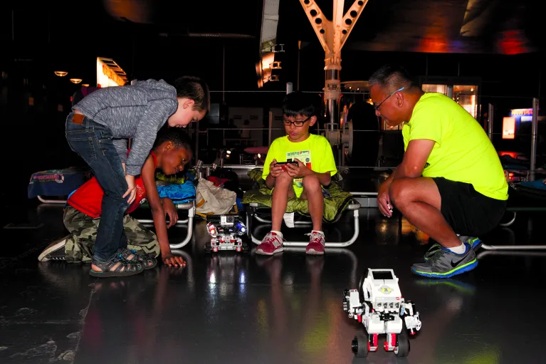 Three children and a chaperon at an overnight. One child is sitting on a cot controlling a robot while the others look on.