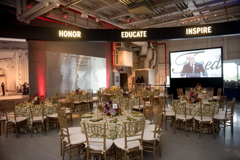 Beautifully set round tables with gold colored chairs. A screen with someone speaking at a podium is in the background.