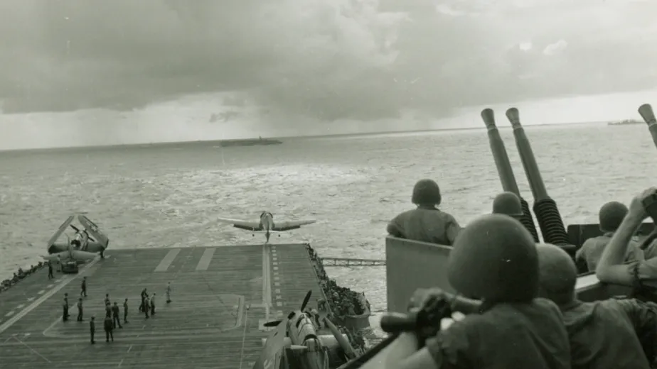 Large battleship moving through the ocean. Planes are lined up on the flight deck, with crew members bustling around.