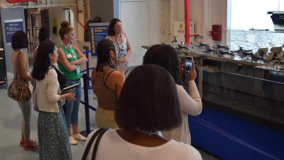 A group of women gathered around, looking closely at a detailed LEGO model of the Intrepid aircraft carrier on display at the Intrepid Museum