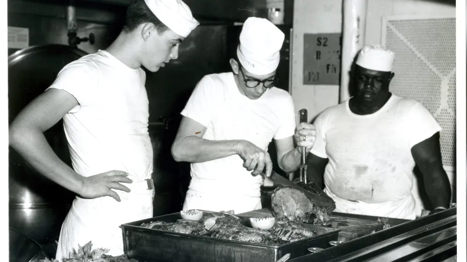 Sailors wearing chef hats carving a piece of meat in the kitchen aboard the Intrepid Museum.